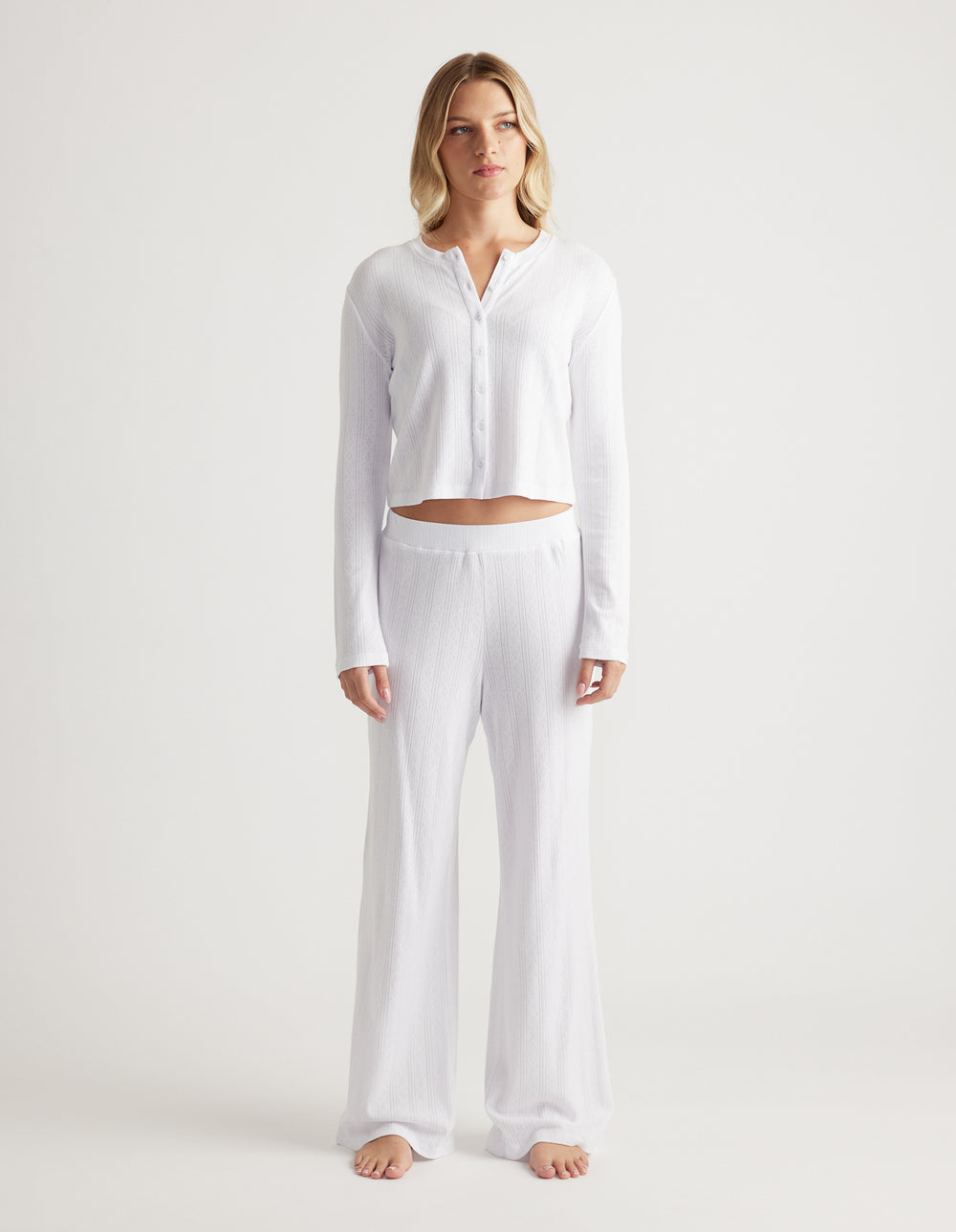 Woman wearing soft white sleep pointelle wide leg pants with elastic waistband and matching long sleeve top with buttons