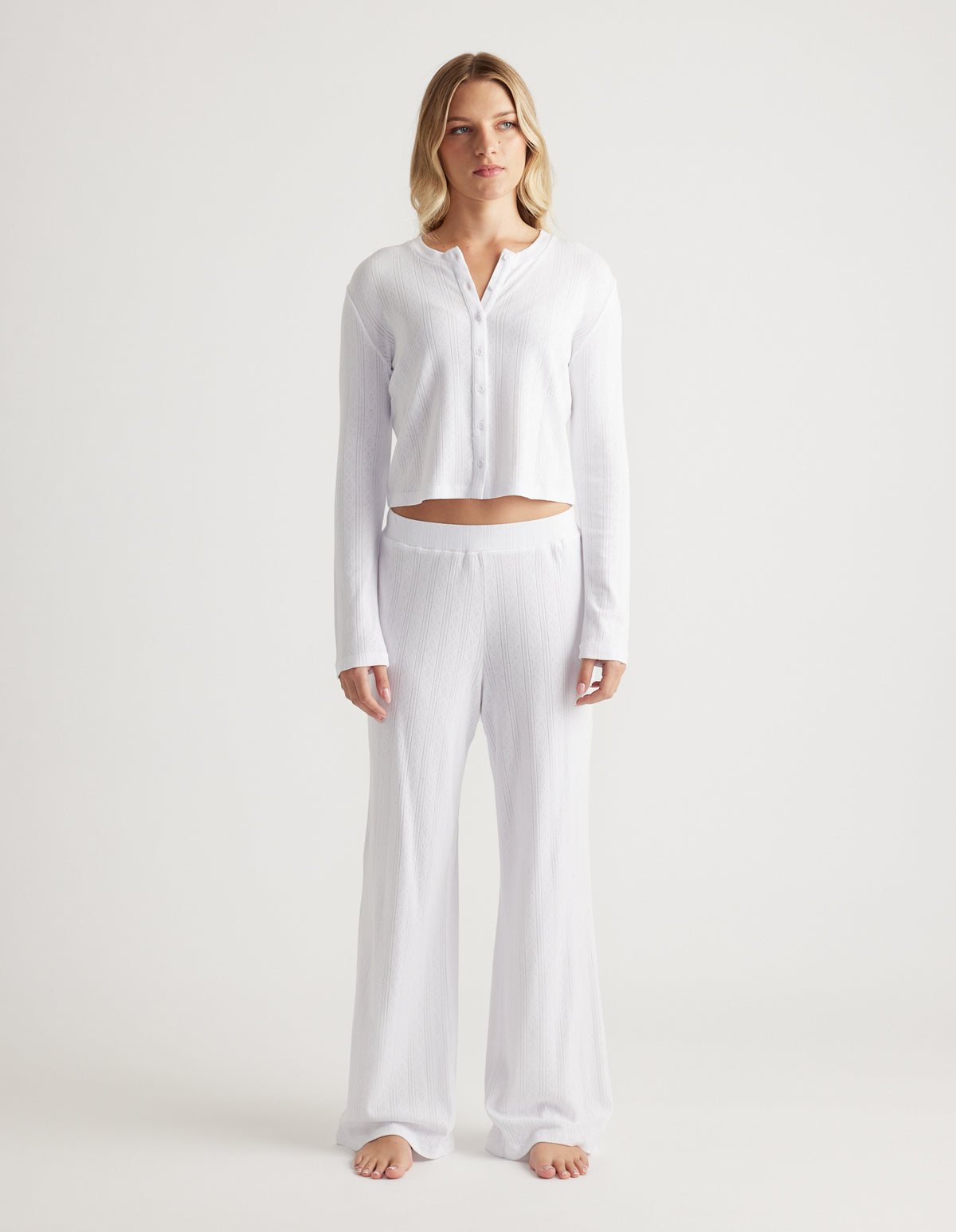 Woman wearing soft white sleep pointelle wide leg pants with elastic waistband and matching long sleeve top with buttons
