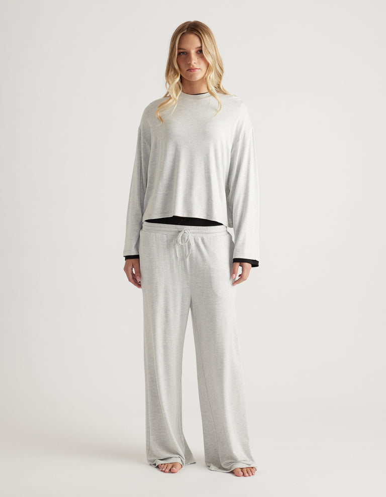 Woman wearing a light grey sleep jersey long sleeve top with matching wide-leg lounge pants against a plain backdrop