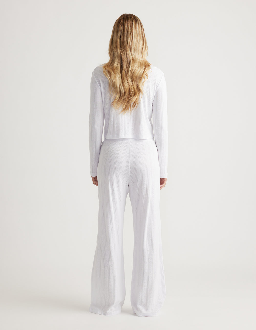 Woman with long wavy blonde hair wearing white wide-leg ribbed pants and matching long-sleeve top viewed from behind