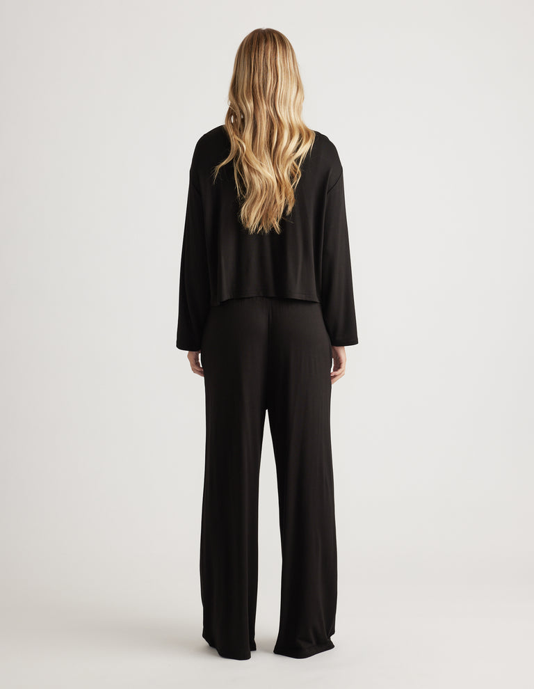 Woman wearing relaxed black wide-leg sleep pants paired with a loose long sleeve top, back view in soft jersey fabric