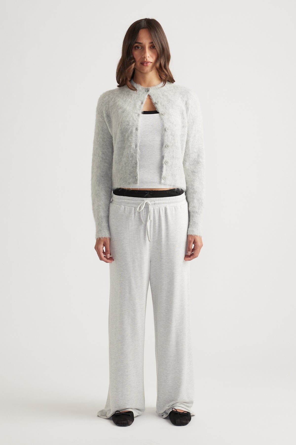 Woman wearing light grey wide leg sleep jersey pants with drawstring waist and a soft fuzzy buttoned cardigan over a white tank top