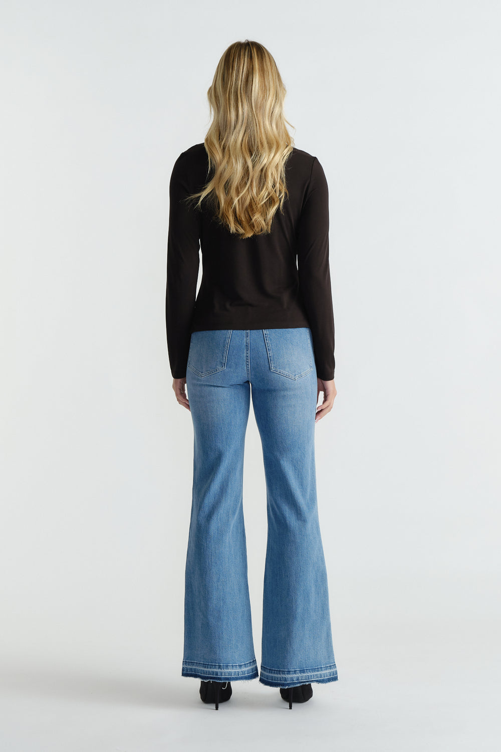 Back view of woman wearing black long-sleeve top paired with high-waisted light blue flared jeans and black heels