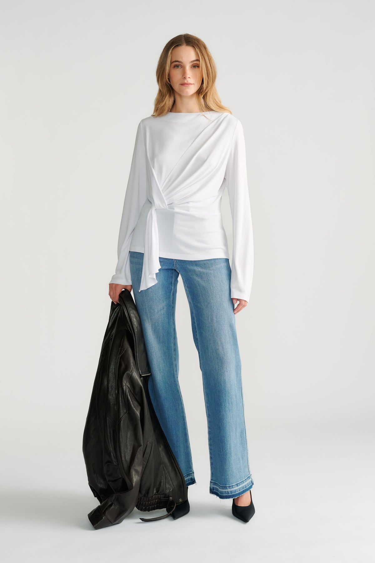 Woman wearing a white long-sleeve top with a front tie detail paired with straight-leg light wash jeans and black heels.