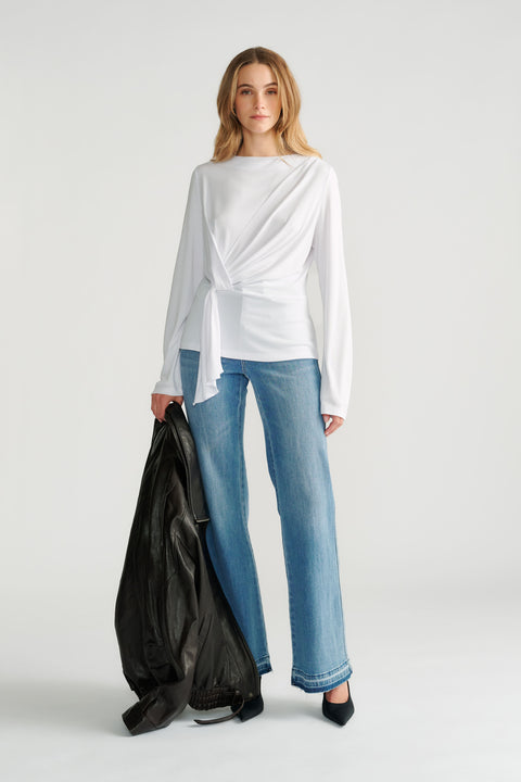 Woman wearing a white long-sleeve top with a front tie detail paired with straight-leg light wash jeans and black heels.