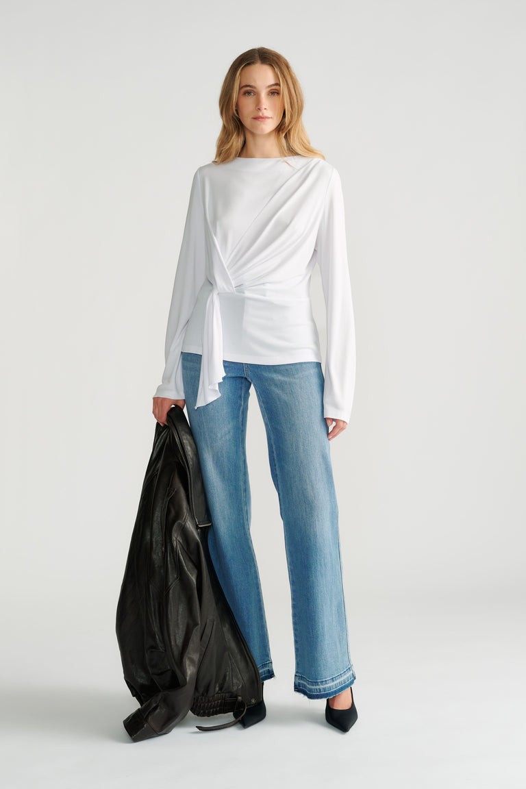 Woman wearing a white long-sleeve top with a front tie detail paired with straight-leg light wash jeans and black heels.