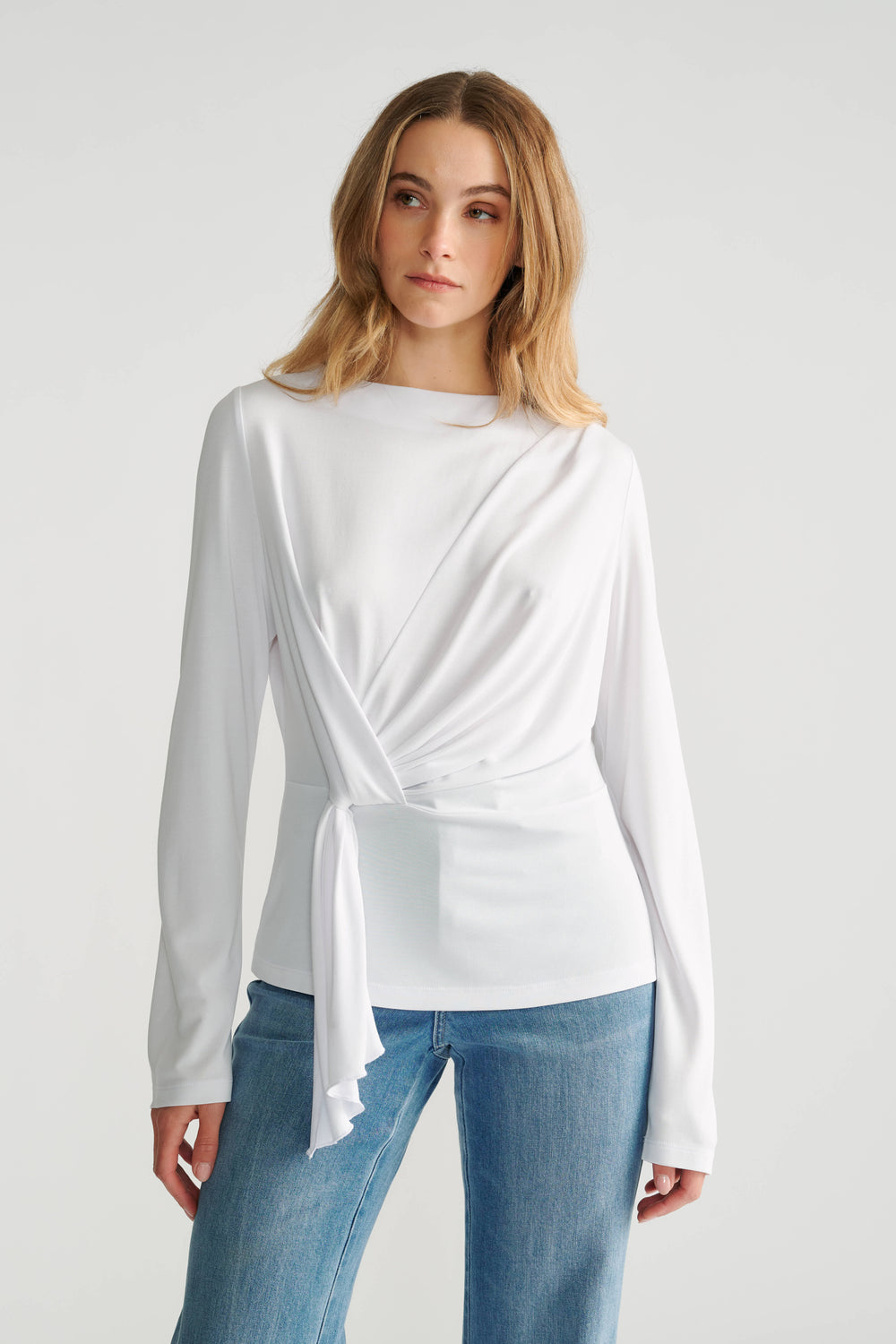 Woman wearing a white long-sleeve top with side tie detail paired with classic mid-blue denim jeans.