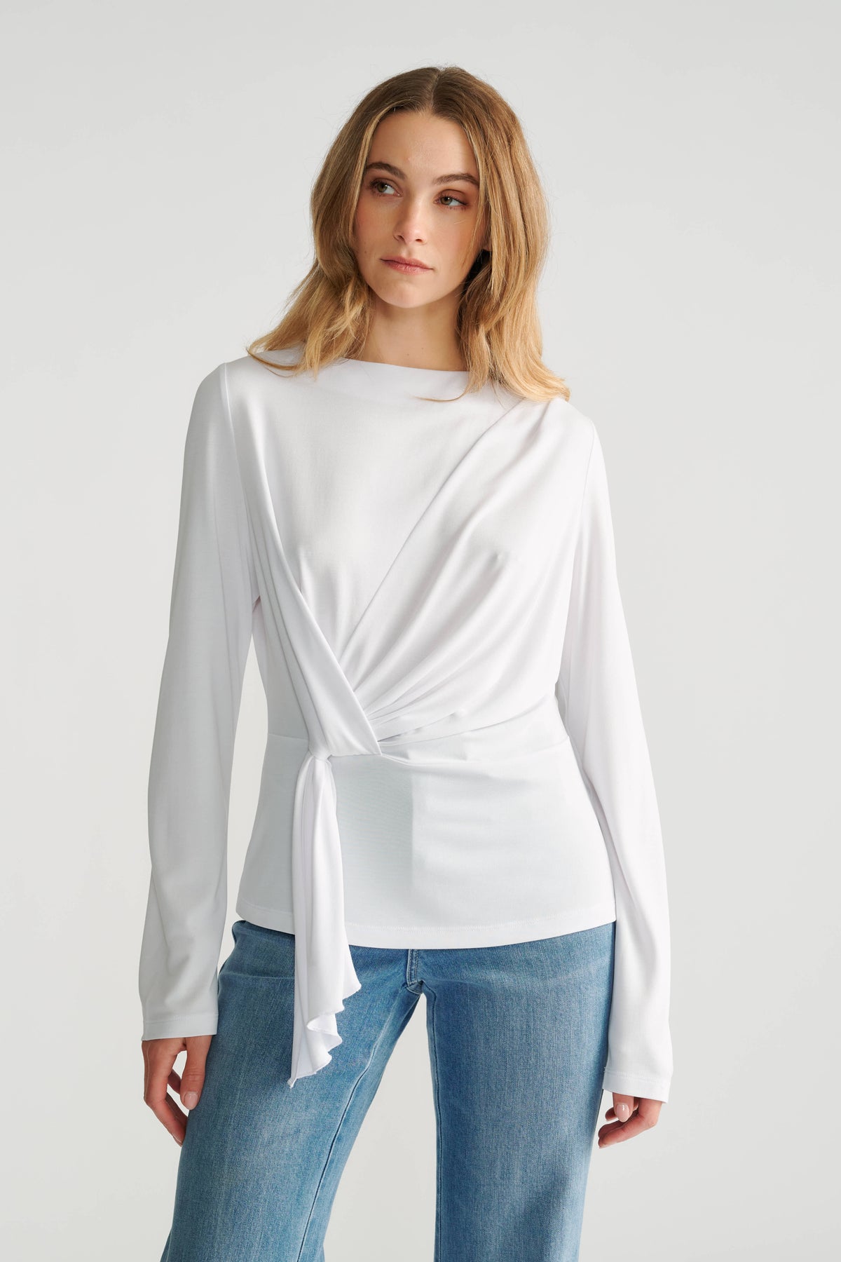 Woman wearing a white long-sleeve top with side tie detail paired with classic mid-blue denim jeans.
