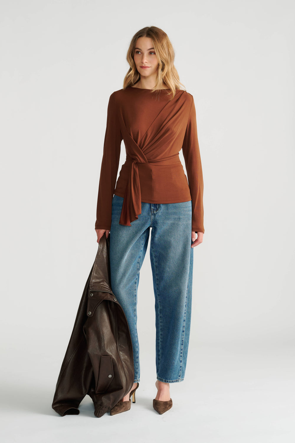 Woman wearing a rust brown long-sleeve top with a wrap tie detail paired with relaxed fit blue jeans and brown heels.