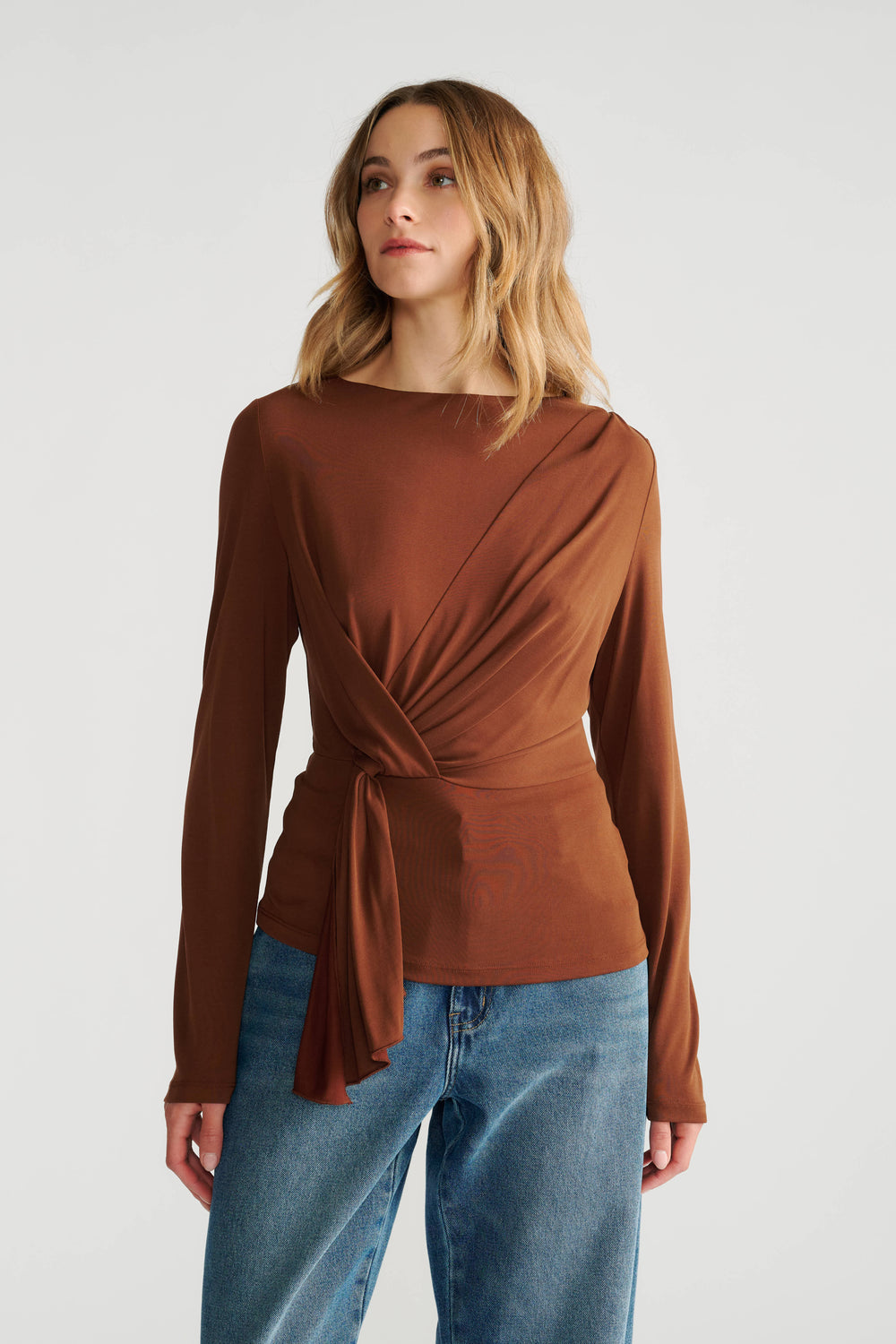 Woman wearing a rich brown long-sleeve top with a side tie detail paired with relaxed blue jeans.
