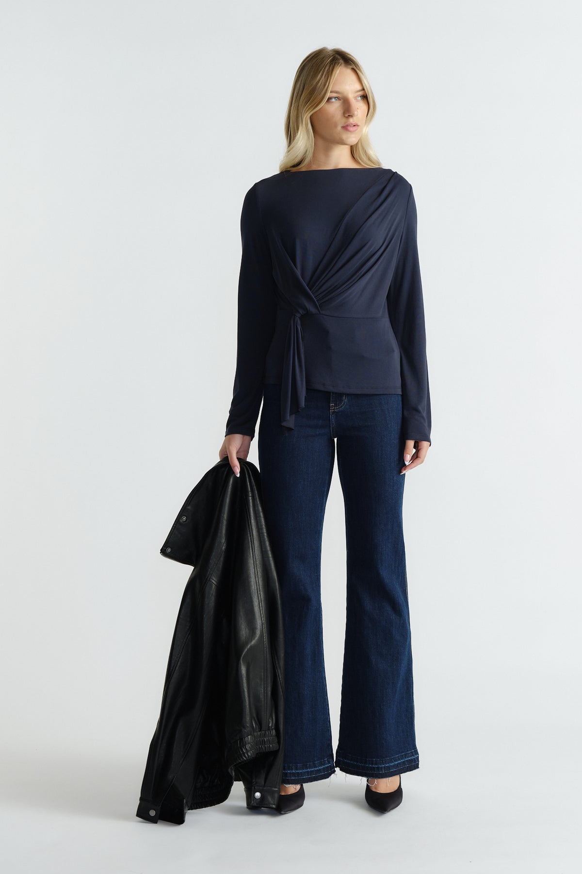 Woman wearing a navy long-sleeve top with side tie detail paired with dark flared jeans and holding a black jacket
