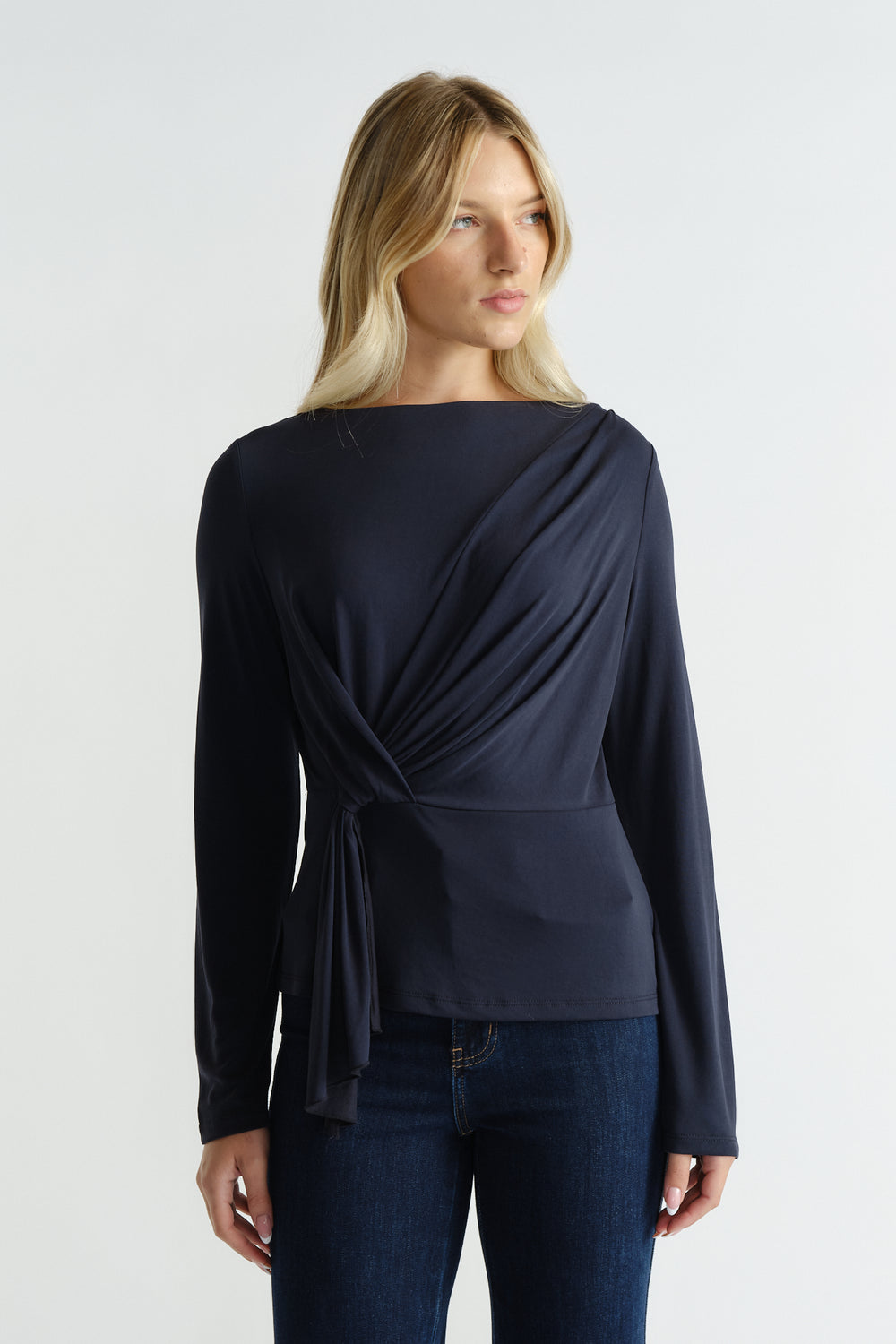 Woman wearing a navy long-sleeve top with a gathered tie detail at the waist, paired with dark denim jeans