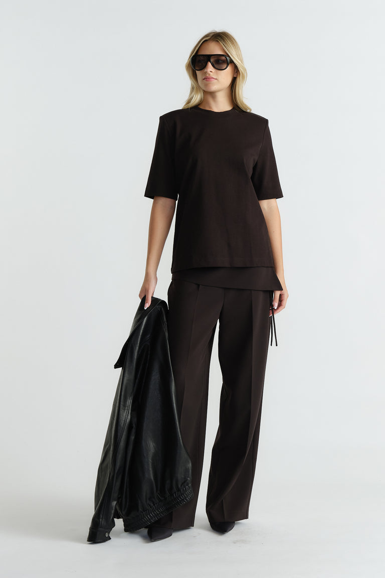Woman wearing a structured black tee with shoulder pads paired with loose wide-leg trousers holding a leather jacket.