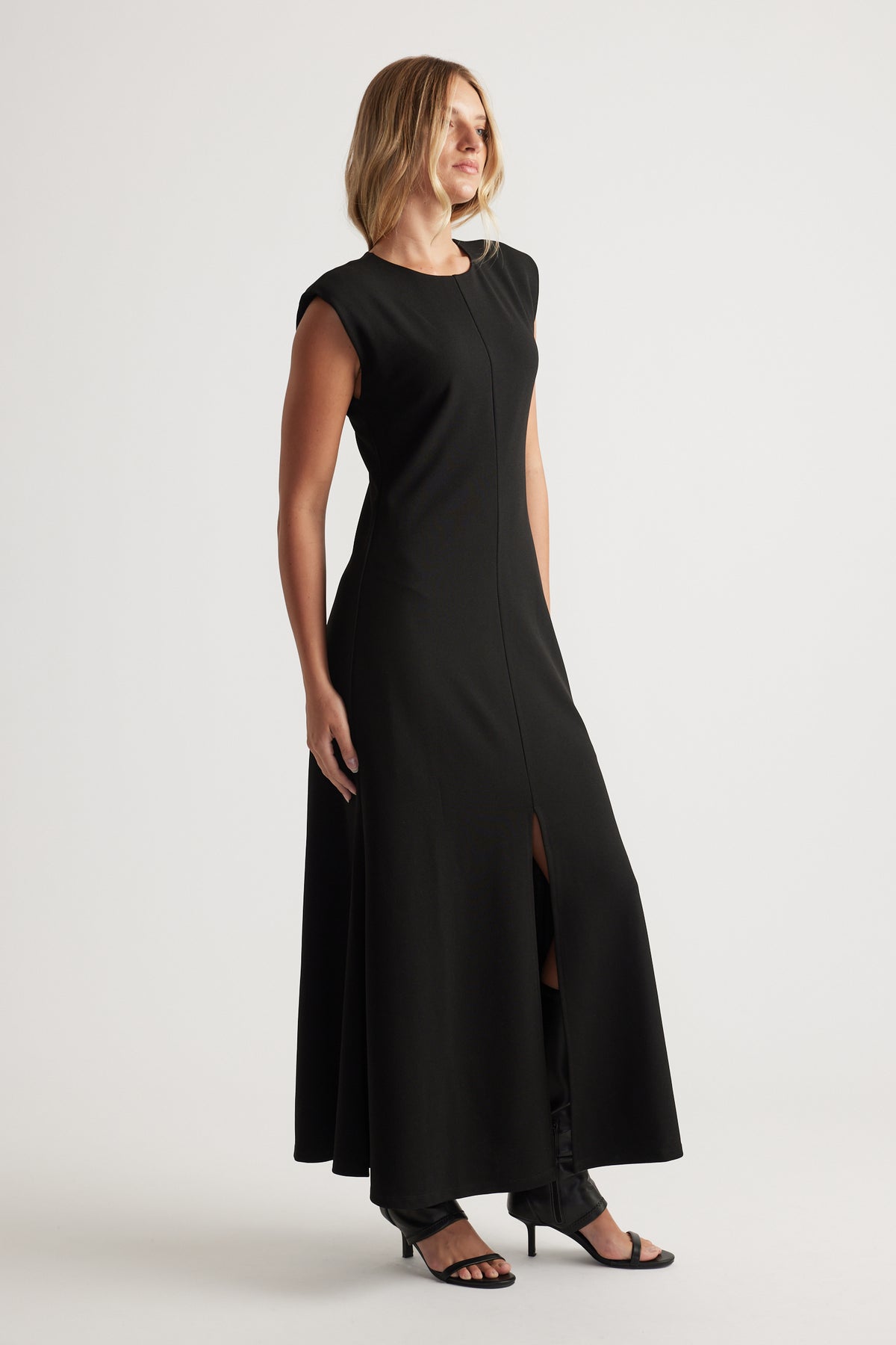 Woman wearing a sleeveless black maxi dress with a front slit and structured shoulder detail paired with black heels