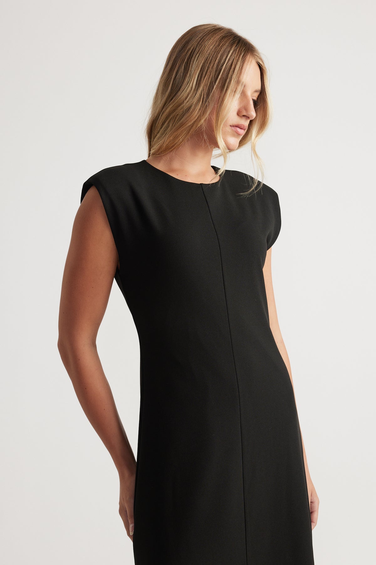 Woman in a sleeveless black dress with a round neck and structured shoulders looking down against a plain background