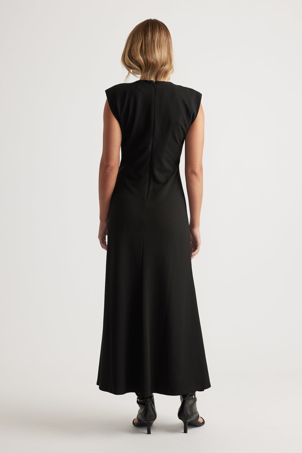 Woman seen from behind wearing a sleeveless black maxi dress with structured shoulders and black heels