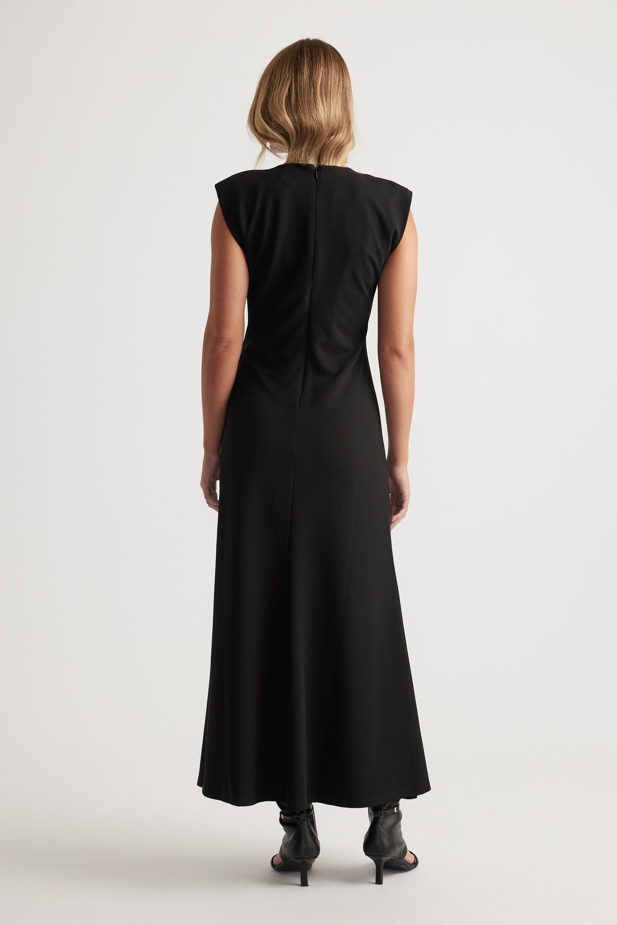 Woman seen from behind wearing a sleeveless black maxi dress with structured shoulders and black heels