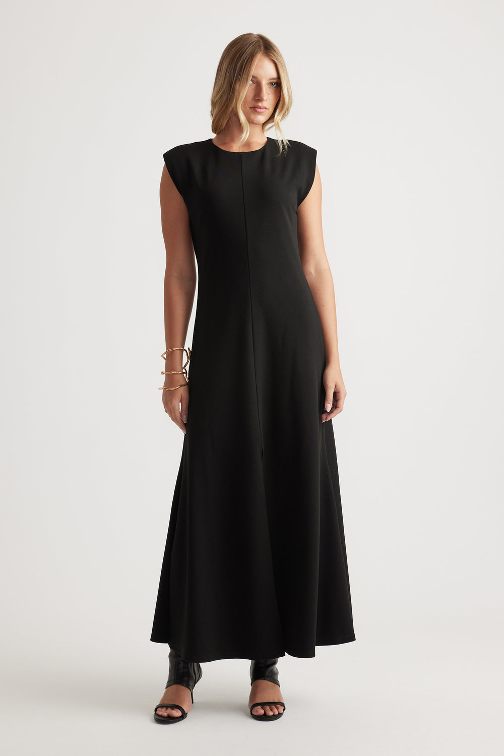 Woman wearing a sleeveless black maxi dress with a round neckline, styled with open-toe black heels and bracelets.