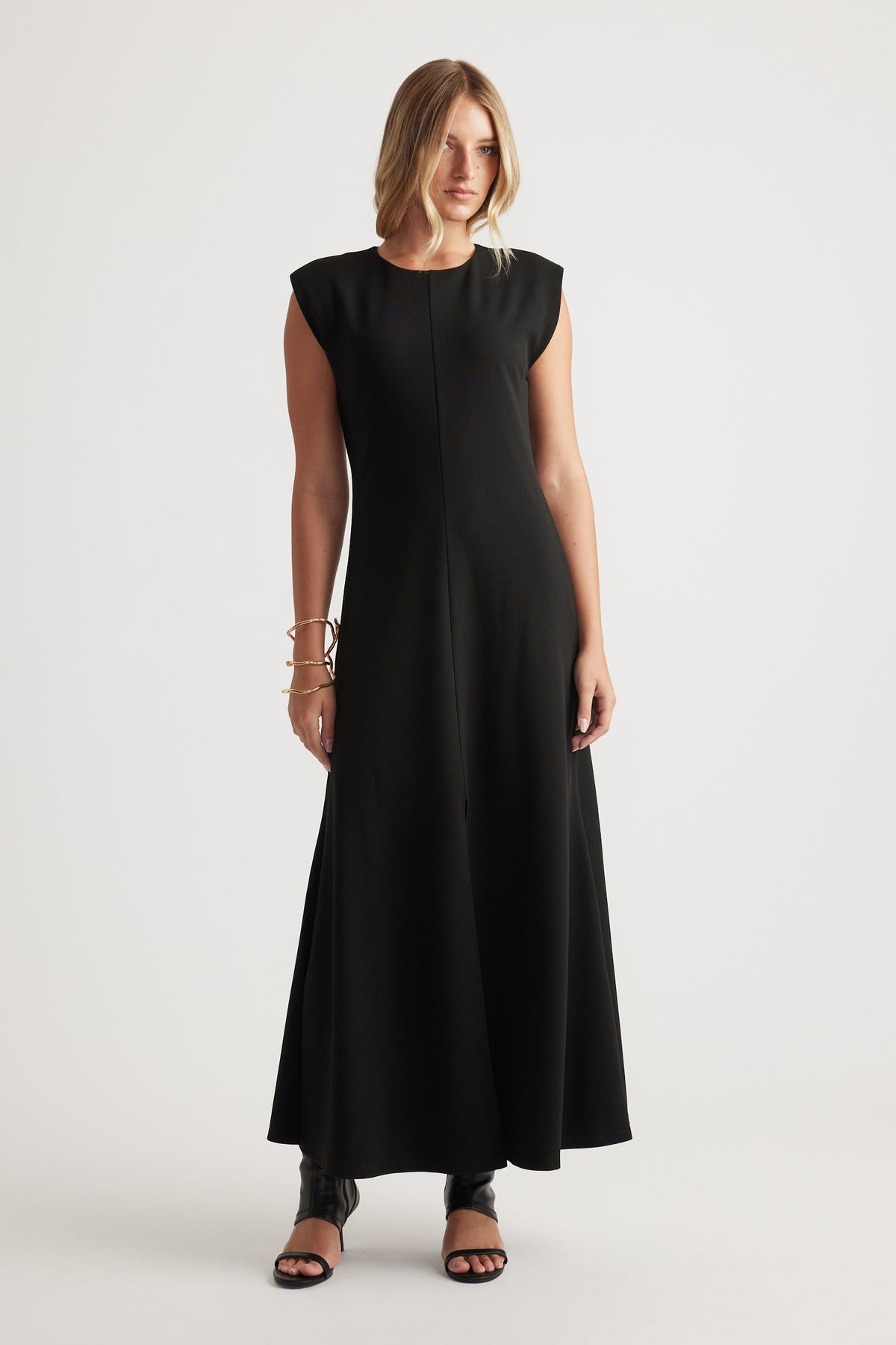 Woman wearing a sleeveless black maxi dress with a round neckline, styled with open-toe black heels and bracelets.