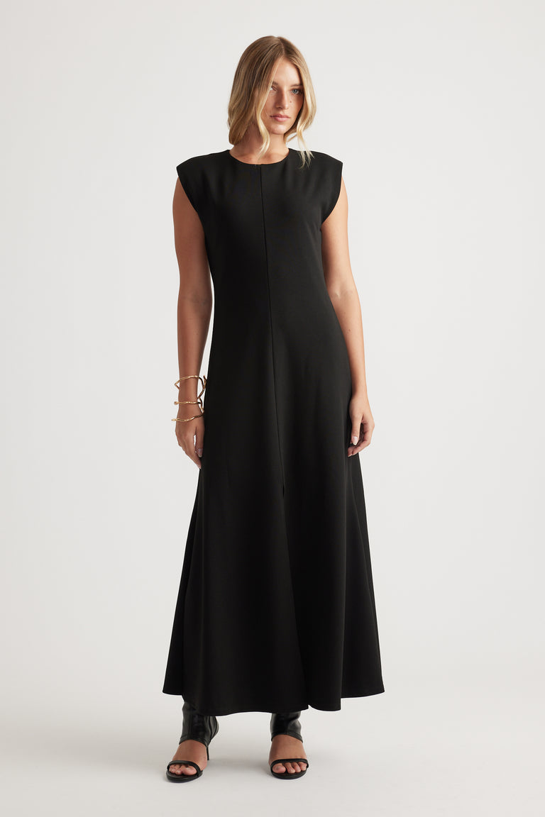 Woman wearing a sleeveless black maxi dress with a round neckline, styled with open-toe black heels and bracelets.