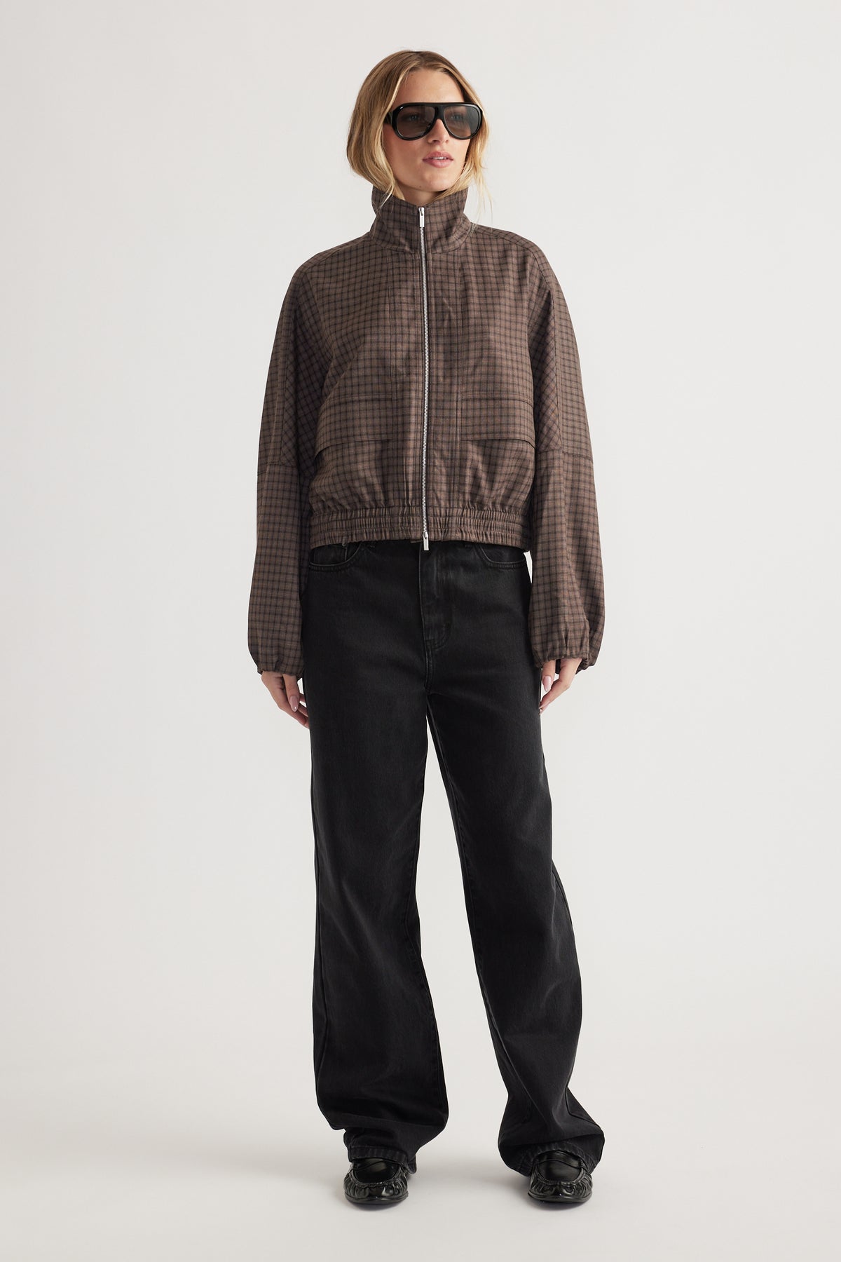 Woman wearing a brown check zip-up jacket with elasticated cuffs and hem paired with loose black pants and sunglasses