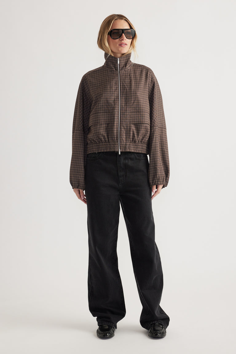 Woman wearing a brown check zip-up jacket with elasticated cuffs and hem paired with loose black pants and sunglasses