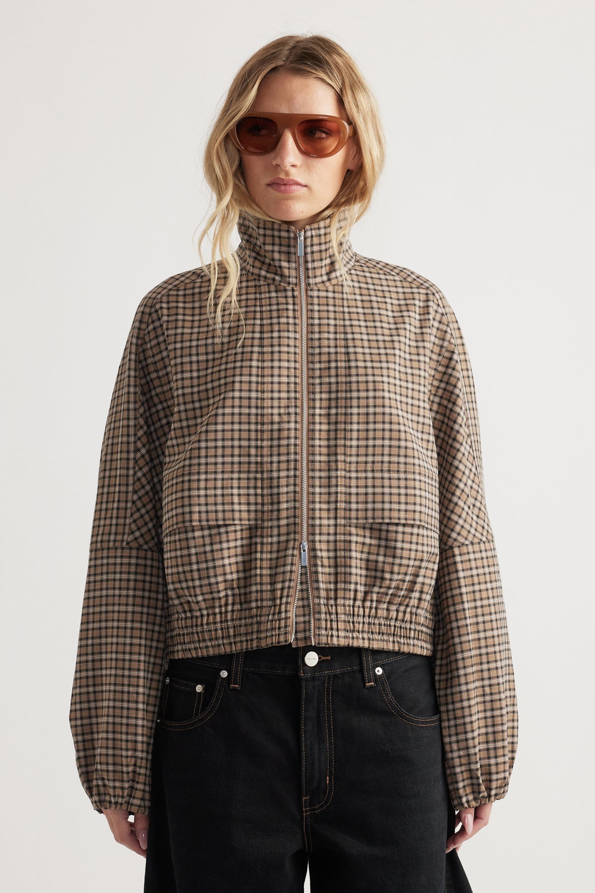 Woman wearing a zip-up check jacket with elasticated cuffs and hem paired with black high-waisted pants and sunglasses