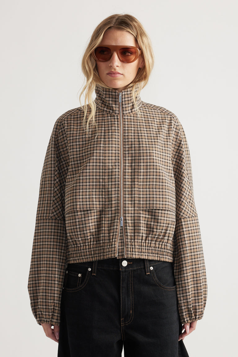 Woman wearing a zip-up check jacket with elasticated cuffs and hem paired with black high-waisted pants and sunglasses