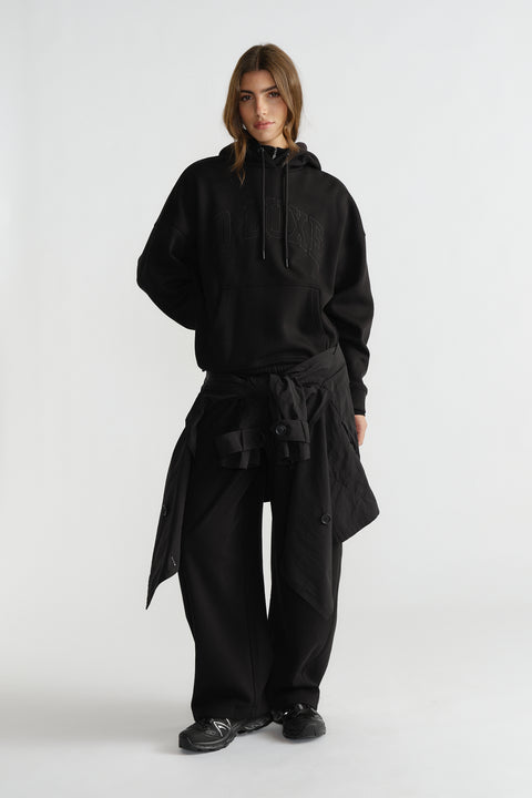 Woman wearing relaxed black hoodie with subtle logo, wide-leg pants, and a lightweight jacket tied at the waist