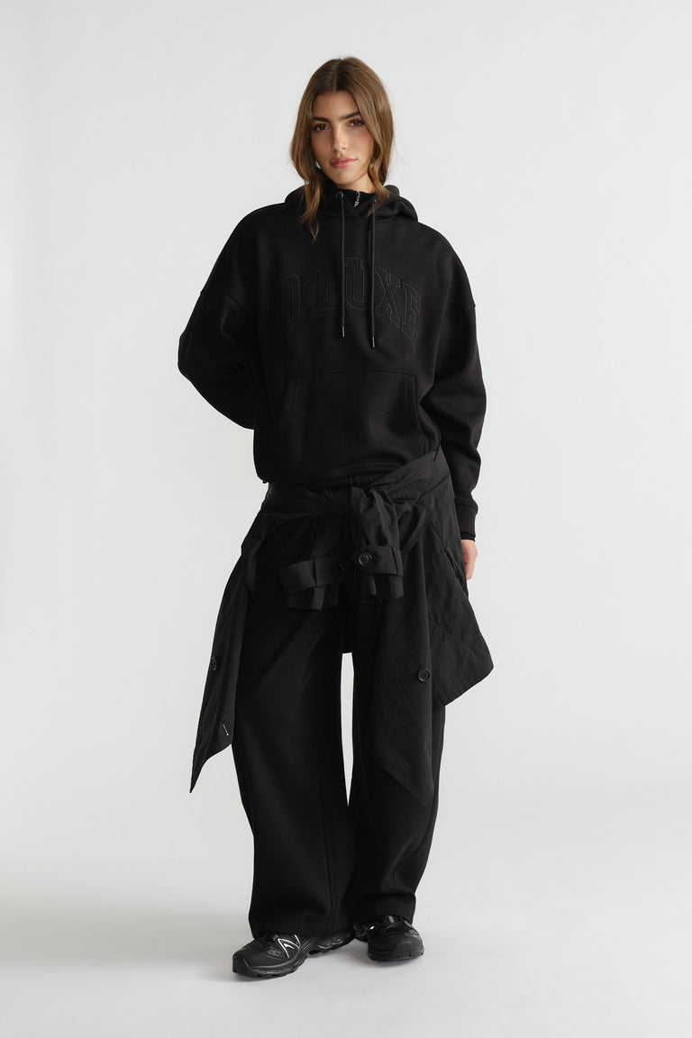 Woman wearing relaxed black hoodie with subtle logo, wide-leg pants, and a lightweight jacket tied at the waist