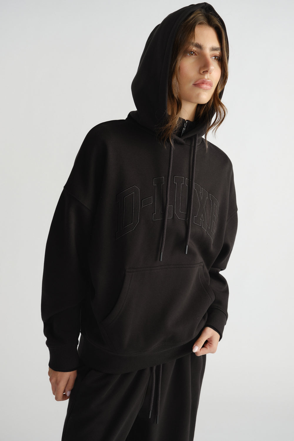 Woman wearing a relaxed black hoodie with subtle embossed "D-LUXE" lettering, featuring a front pocket and drawstring hood