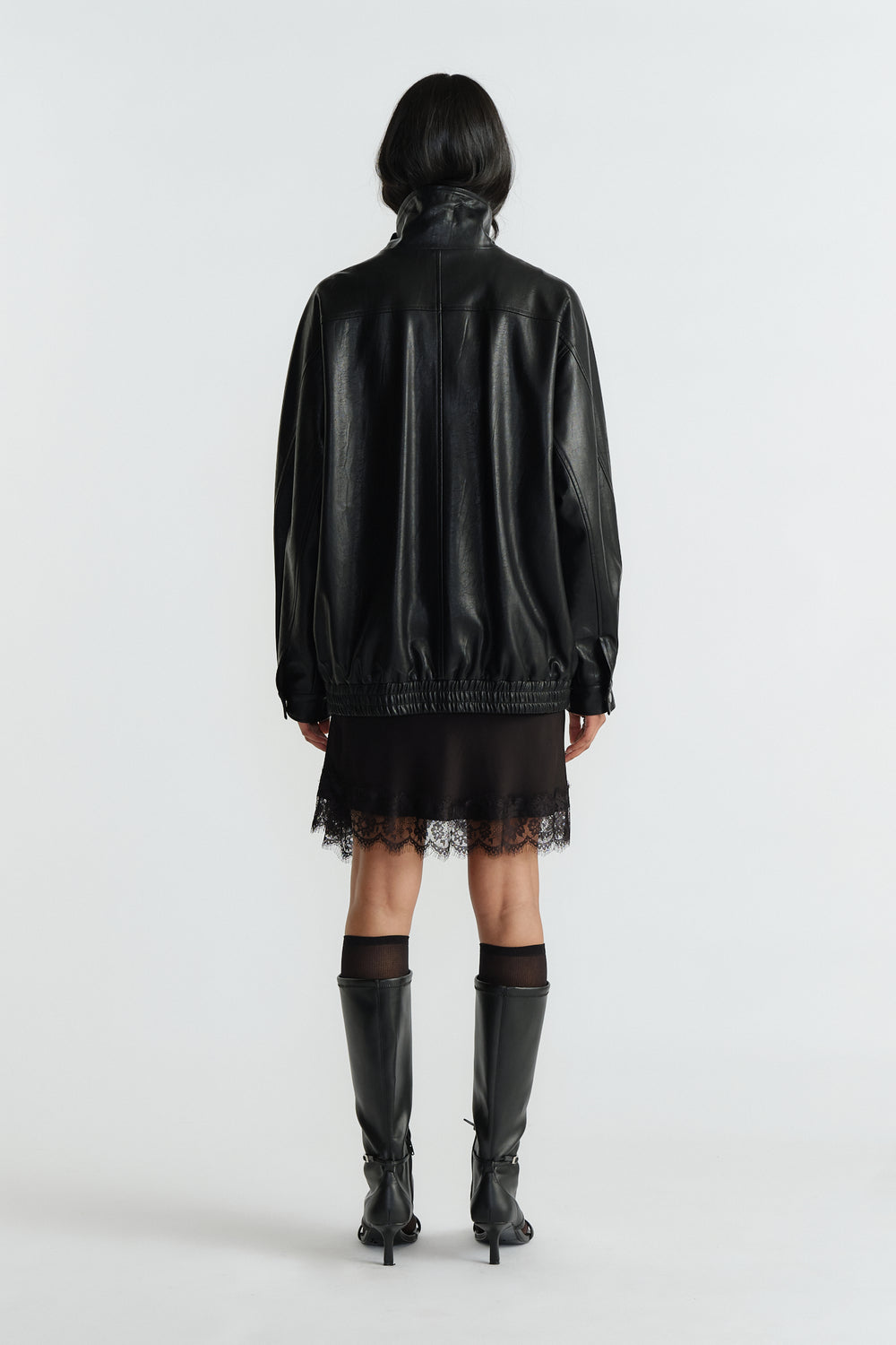 Back view of woman wearing an oversized black jacket with gathered hem paired with a lace-trimmed black dress and knee boots