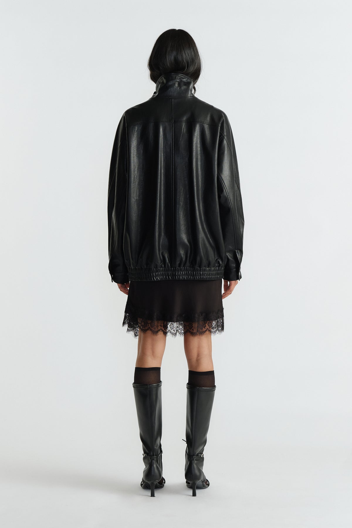 Back view of woman wearing an oversized black jacket with gathered hem paired with a lace-trimmed black dress and knee boots