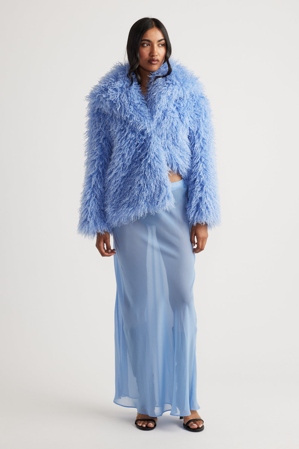 Model wearing a light blue sheer maxi skirt with a voluminous textured blue jacket and black open-toe heels