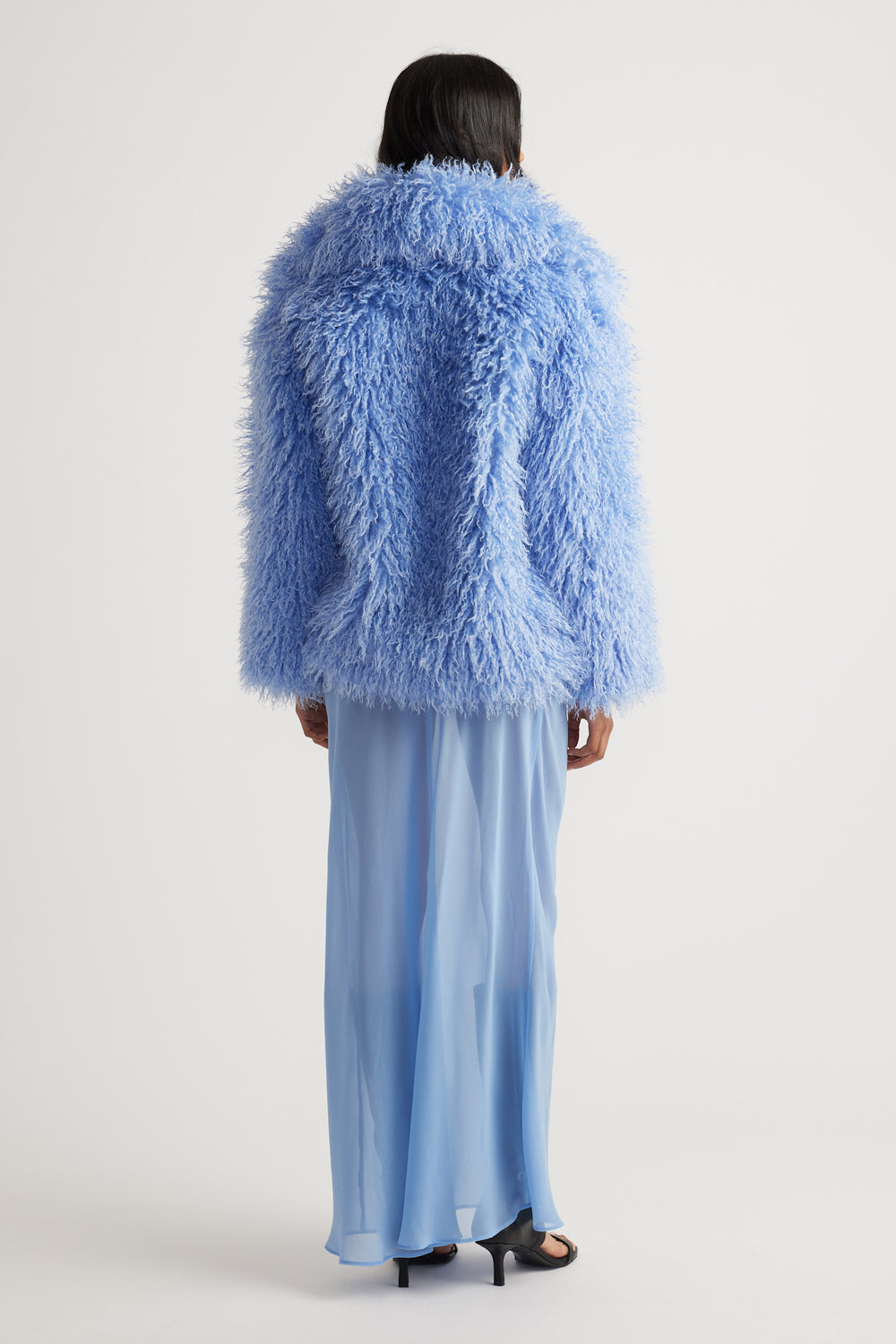 Back view of a light blue textured fluffy jacket paired with a flowing sheer maxi skirt and black heeled sandals
