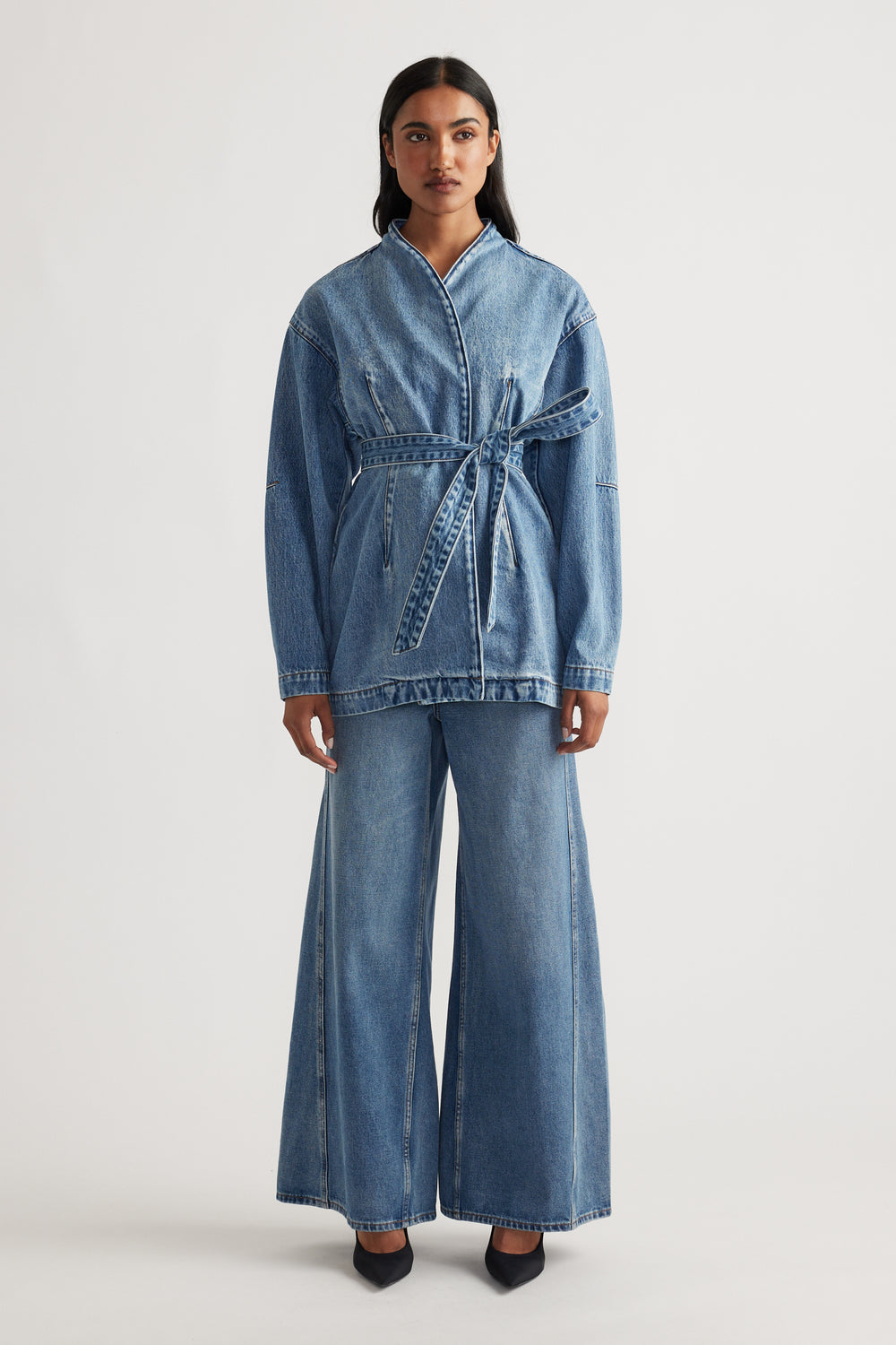 Woman wearing a belted denim jacket paired with high-waisted wide leg blue jeans and black pointed shoes
