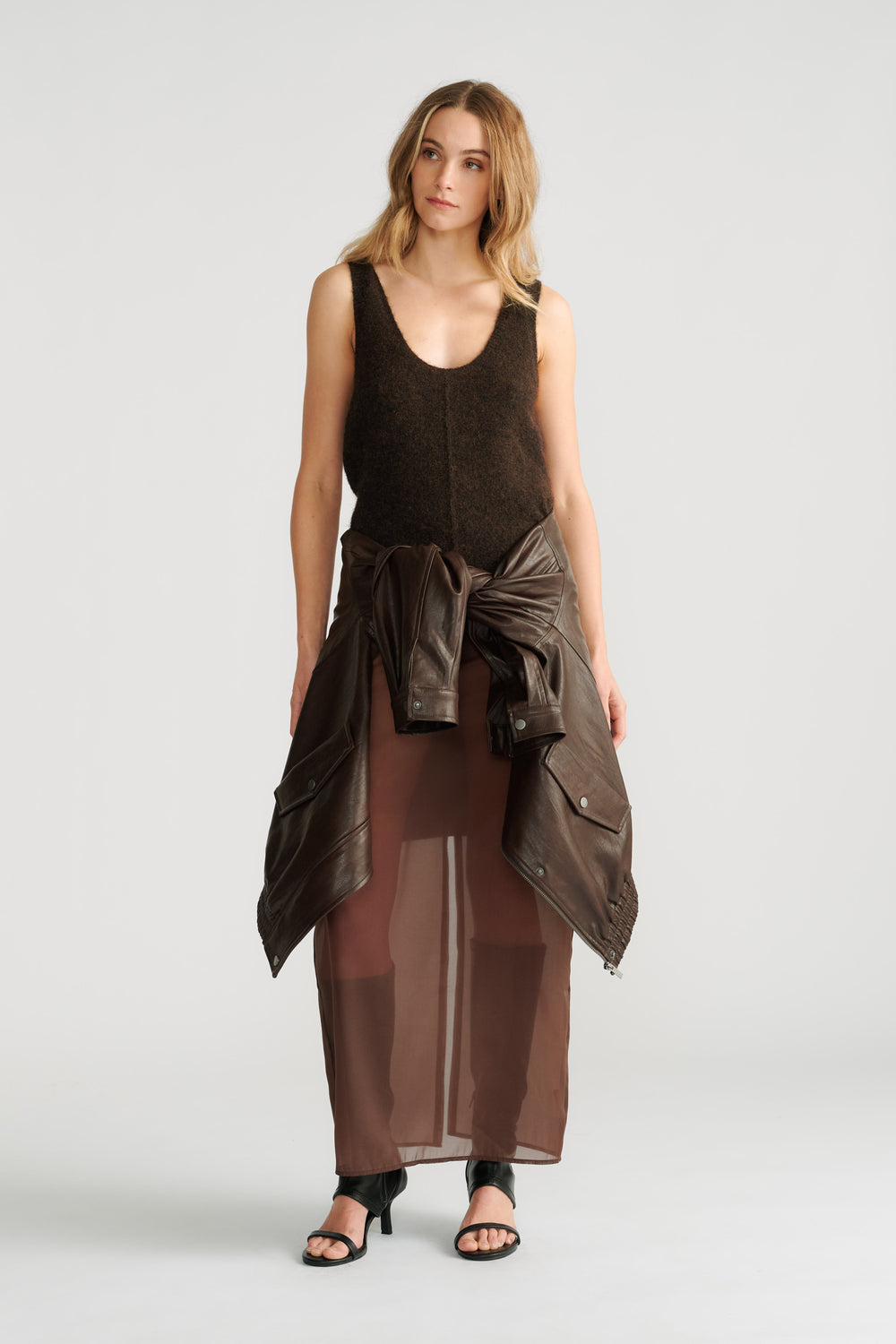 Woman wearing a dark brown fluffy knit tank with a tied leather jacket and sheer brown maxi skirt paired with black heels