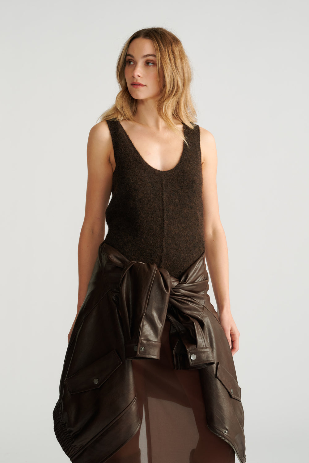 Woman wearing a dark brown textured knit tank paired with a leather jacket tied at the waist and a sheer skirt.