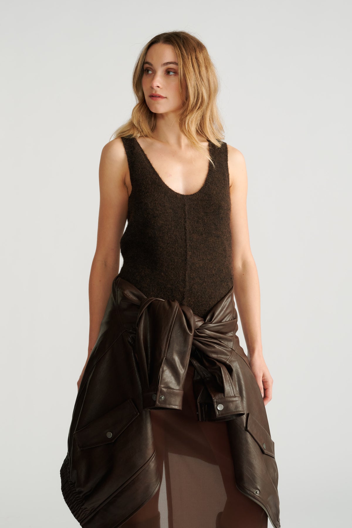 Woman wearing a dark brown textured knit tank paired with a leather jacket tied at the waist and a sheer skirt.