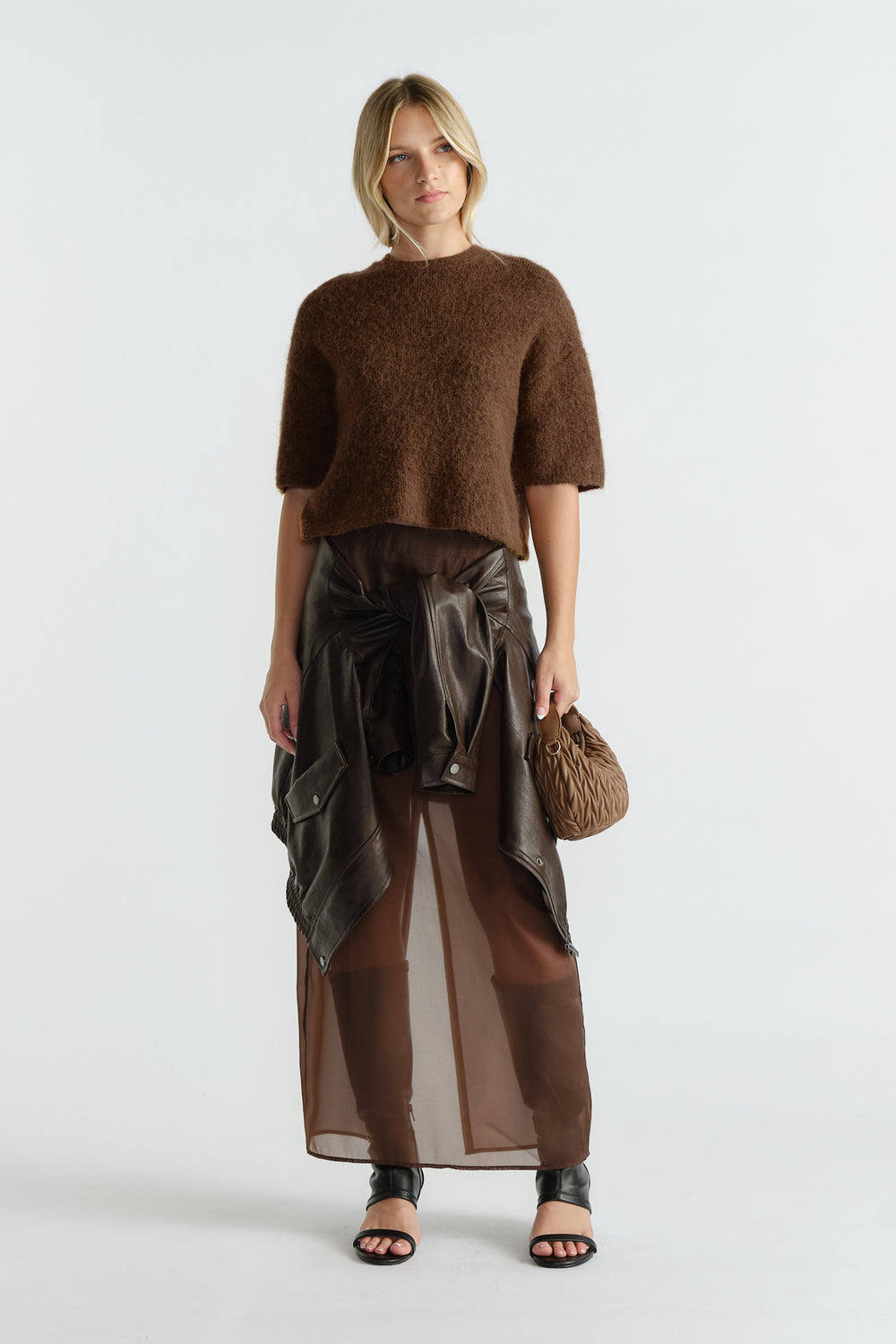 Woman wearing a textured brown knit tee paired with a sheer brown maxi skirt, black sandals, and a tied leather jacket at waist.