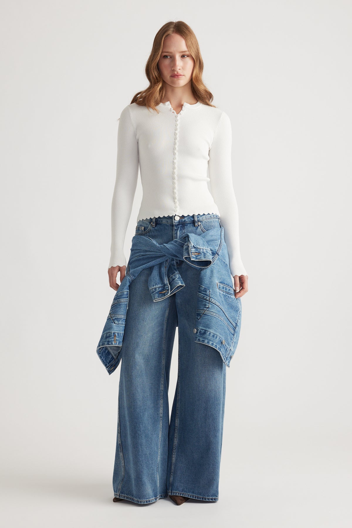 Woman wearing a white scalloped button top paired with wide leg blue jeans and a denim jacket tied at the waist