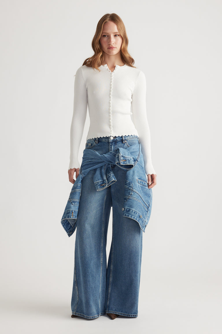 Woman wearing a white scalloped button top paired with wide leg blue jeans and a denim jacket tied at the waist