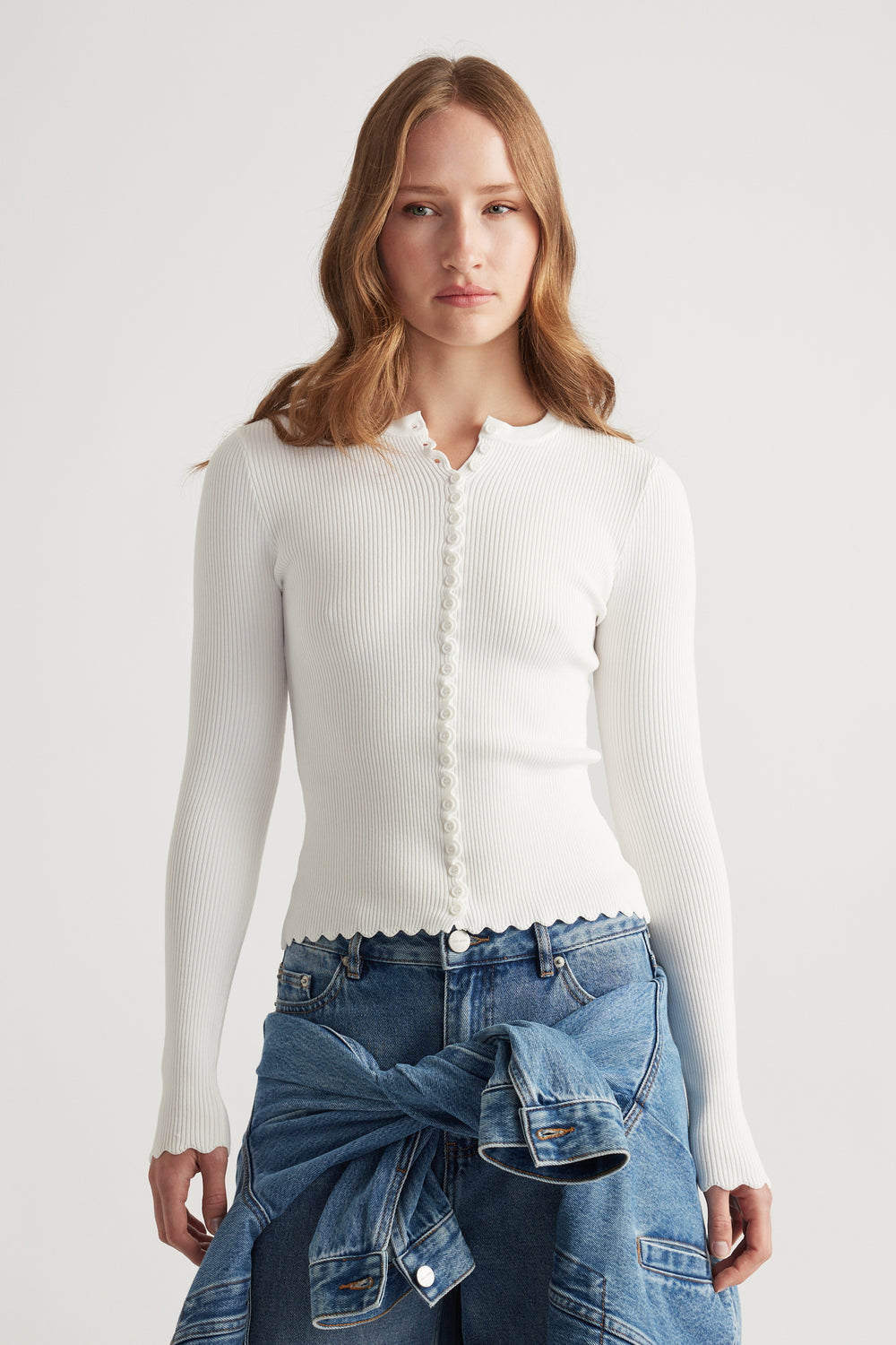 Woman wearing a fitted white scalloped button long sleeve top paired with blue denim jeans and a tied jacket around waist