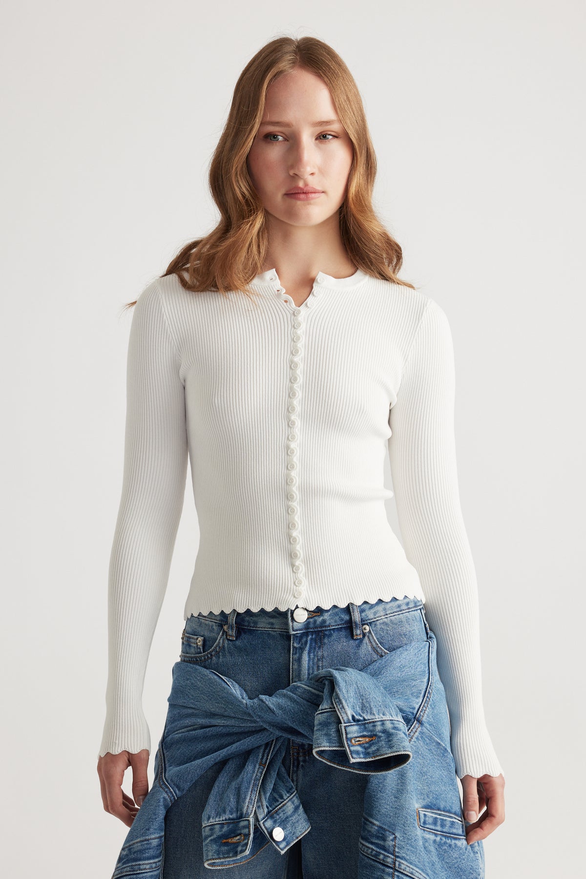 Woman wearing a fitted white scalloped button long sleeve top paired with blue denim jeans and a tied jacket around waist