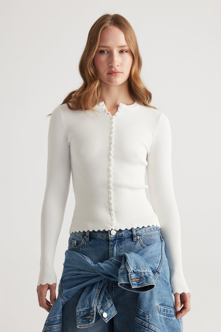 Woman wearing a fitted white scalloped button long sleeve top paired with blue denim jeans and a tied jacket around waist