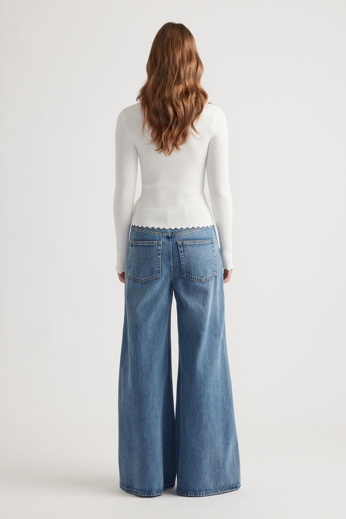 Woman wearing a white scalloped-edge long-sleeve top paired with wide-leg mid-blue denim jeans viewed from the back