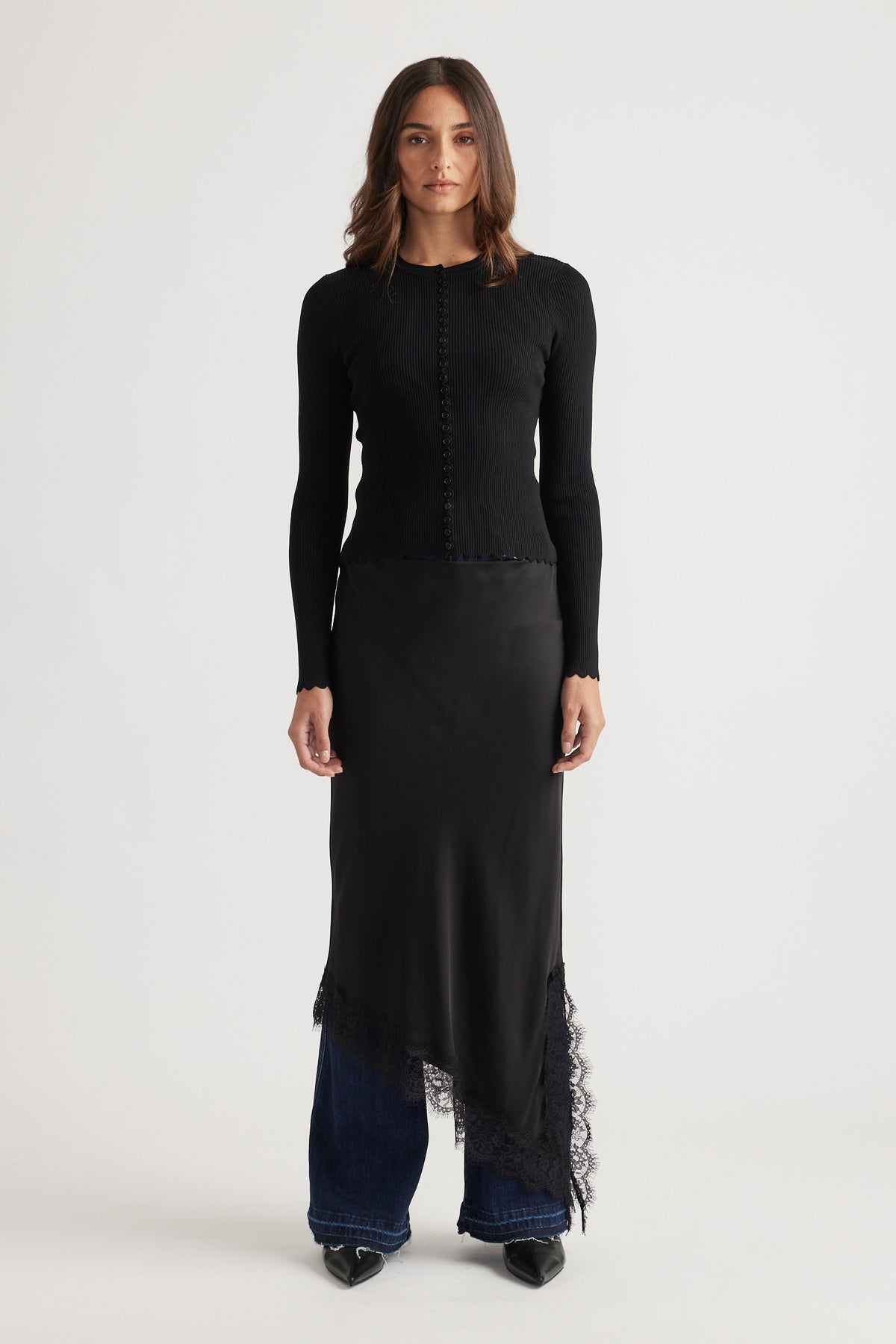 Woman wearing a fitted black top with scalloped button details paired with an asymmetrical lace-trimmed skirt and jeans