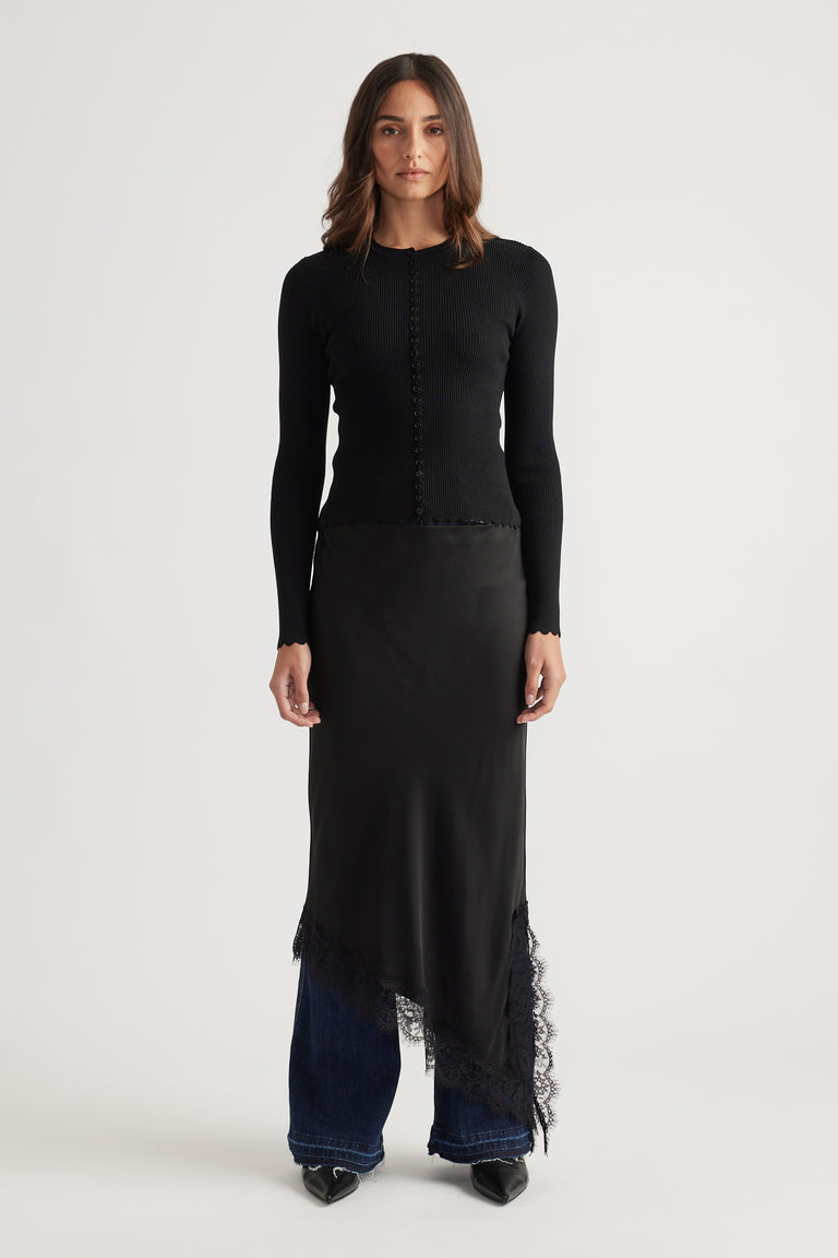 Woman wearing a fitted black top with scalloped button details paired with an asymmetrical lace-trimmed skirt and jeans
