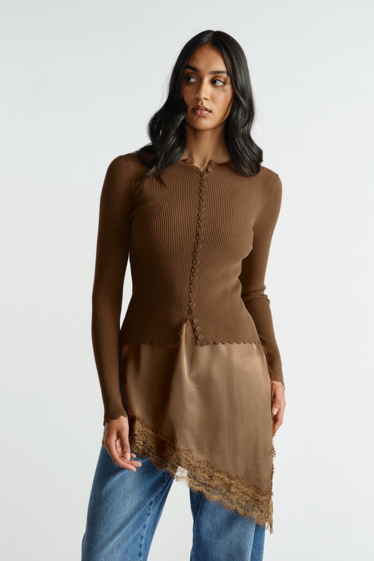 Woman wearing a fitted brown ribbed button-up top with scalloped edges over an asymmetric lace-trimmed layer and jeans