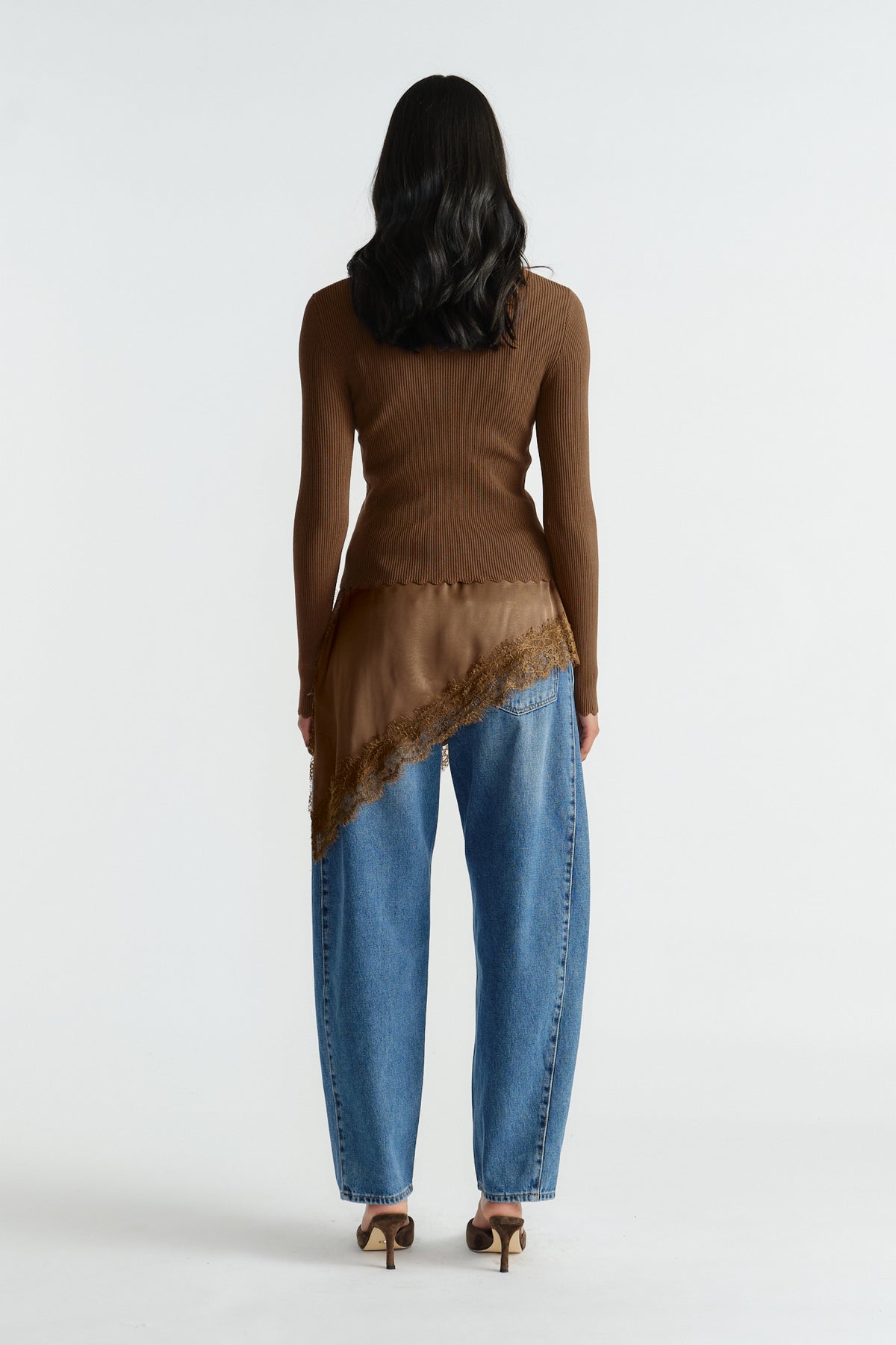 Back view of woman wearing a fitted brown knit top layered with a scalloped asymmetric caramel silk detail and relaxed blue jeans